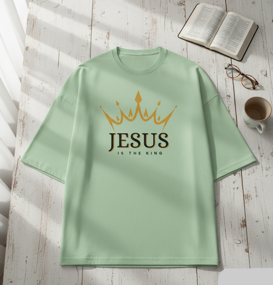 Light green t-shirt with 'Jesus is the King' text on a wooden surface with a book and cup.